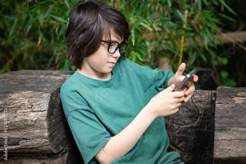 Young teenager boy wearing glasses sitting on eco bench in the park, typing message on his smartphone, child using phone outdoor in summer, social media concept.
