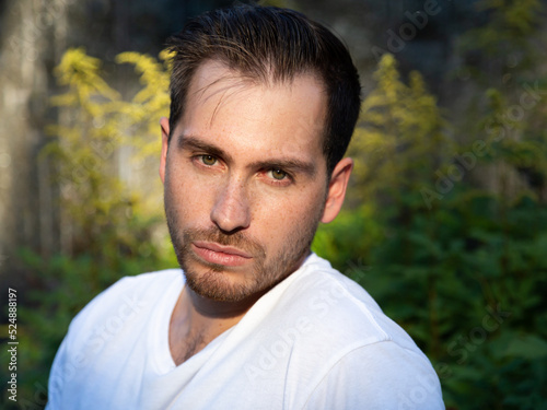 Horizontal closeup of caucasian young man with rugged good looks standing with serious expression in park