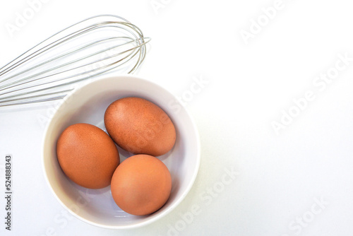 Top view of chickken eggs in a bowl and egg whisks on white background. Copy space.