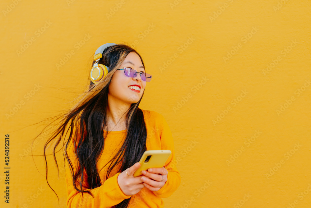 young asian woman wearing sunglasses using her smart phone to listen to music with wireless earphones. girl portrait on yellow background. young asian woman wearing sunglasses using her smart phone to listen to music with wireless earphones. girl portrait on yellow background.