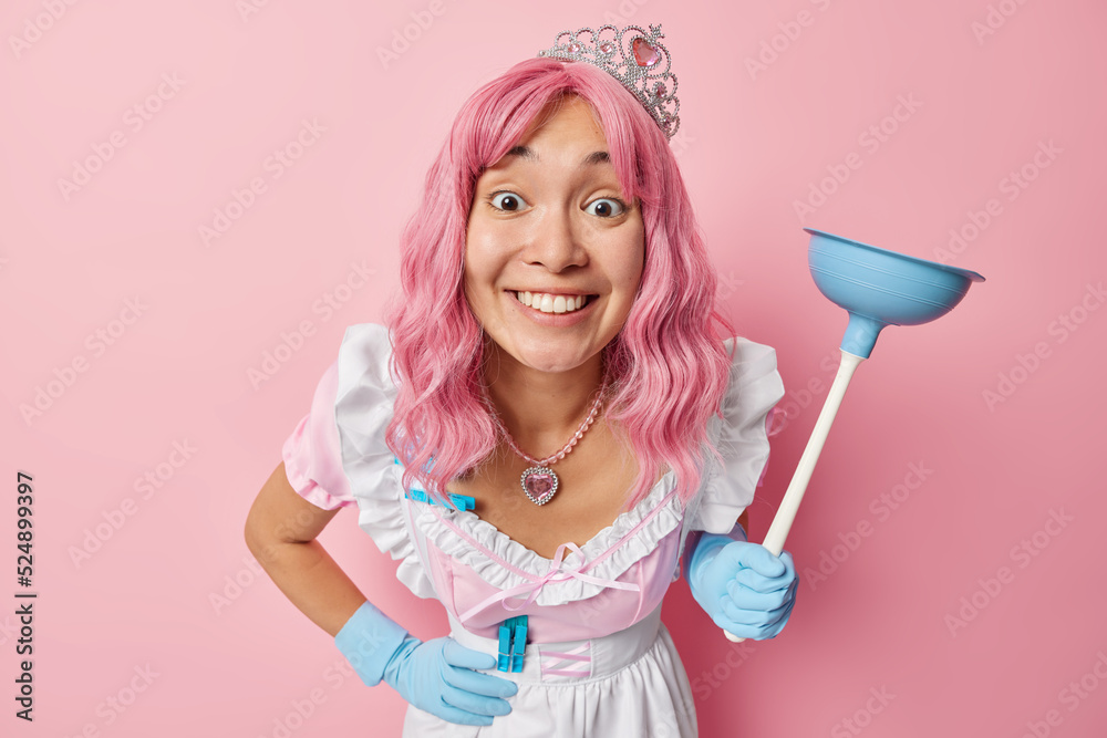 Positive Asian housemaid with pink dyed hair wears crown dress and rubber gloves focused directly at camera holds plunger does house cleaning isolated over pink background. Housekeeping concept Positive Asian housemaid with pink dyed hair wears crown dress and rubber gloves focused directly at camera holds plunger does house cleaning isolated over pink background. Housekeeping concept