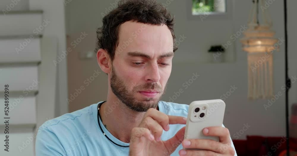Handsome Caucasian man in casual blue t-shirt relaxing in the home interior, using smartphone, types text while messaging, checking news feed and social media content. Online communication Handsome Caucasian man in casual blue t-shirt relaxing in the home interior, using smartphone, types text while messaging, checking news feed and social media content. Online communication