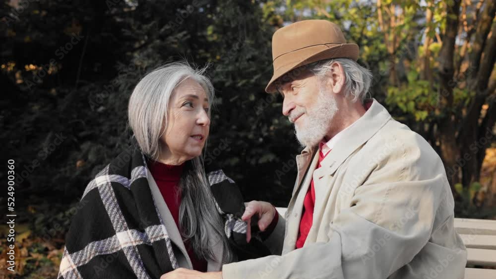 Middle aged spouses enjoying time together in beautiful autumn park. Happy old mature retired couple sitting on a park bench, talking and smiling to each other. Romantic date.