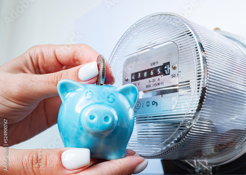 Coin and piggy bank in woman's hands near electricity meter. Symbolic image of energy costs and electricity prices, saving the household budget on paying utility bills.