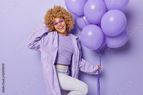 Cheerful curly haired woman poses with bunch of inflated balloons wears stylish clothes of one color celebrates special occasion comes on party isolated over purple background. Holiday concept