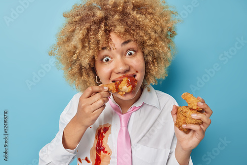 Positive surprised curly haired woman eats delicious nuggets with ketchup enjoys eating junk food wears white formal shirt with tie isolated over blue background. Unhealthy nutrition concept
