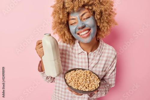 Positive carefree woman with curly hair smiles toothily keeps eyes closed from pleasure applies beauty clay mask holds bottle of milk and cornflakes dressed in nightwear isolated over pink background