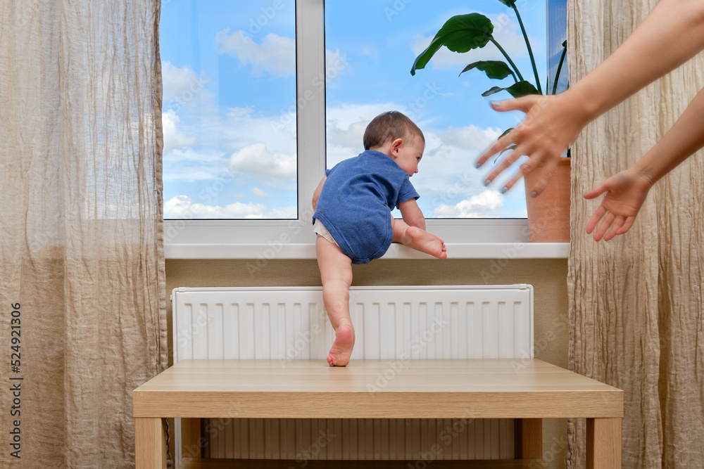 Toddler baby climbs to the window and the hands of a frightened mother. Child crawls to the window holding on to the furniture in the home living room. Kid age one year Toddler baby climbs to the window and the hands of a frightened mother. Child crawls to the window holding on to the furniture in the home living room. Kid age one year