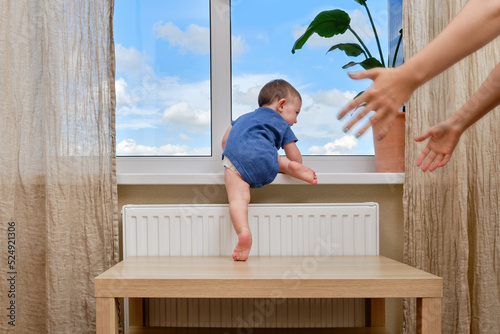 Toddler baby climbs to the window and the hands of a frightened mother. Child crawls to the window holding on to the furniture in the home living room. Kid age one year photo