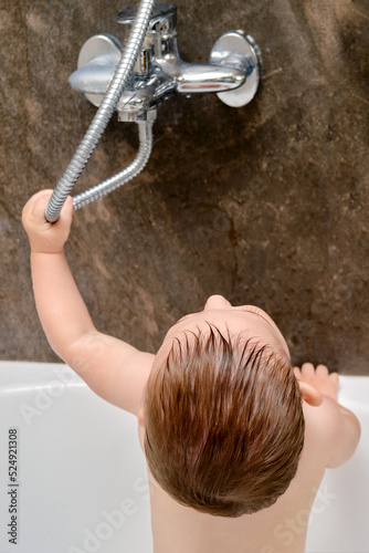 Toddler baby boy is playing with a faucet and a shower while standing in the bathtub. A child dabbles with water while bathing in the bathroom. Kid age one year photo