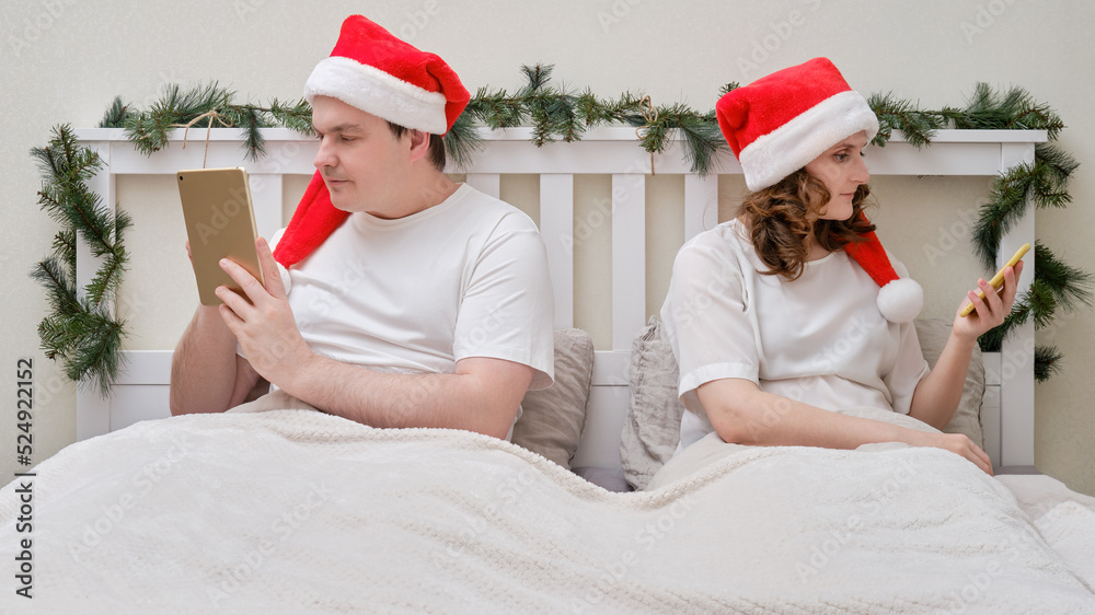 Fototapeta premium A woman and a man are looking at a tablet facing away from each other on a bed decorated for Christmas on New Year Eve, couple in a home bedroom