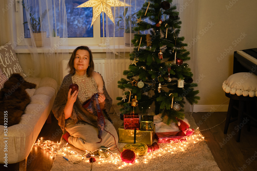 An adult woman knits clothes by the Christmas tree in the evening living room. Hobbies and needlework with wool yarn on New Year Eve