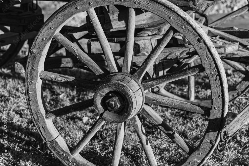 This is a digitally enhanced, monochrome closeup image of a buckboard wagon wheel.  The evening light and shadow accentuates the texture of the weathered wood and antique iron.