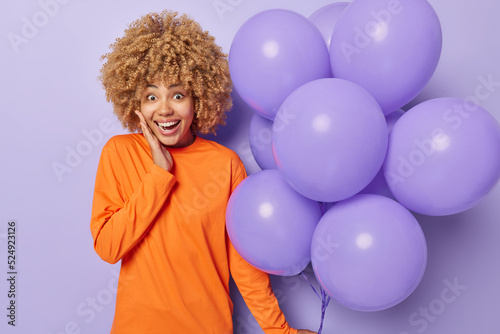 Happy surprised young woman with curly hair feels positive receives congratulations on birthday dressed in casual orange jumper holds bunch of inflated balloons isolated over purple background