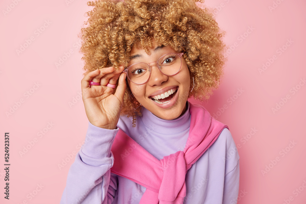 Portrait of cheerful woman with curly hair smiles broadly keeps hand on rim of spectacles notices something pleasant feels delighted dressed in casual clothing isolated over pink background. Portrait of cheerful woman with curly hair smiles broadly keeps hand on rim of spectacles notices something pleasant feels delighted dressed in casual clothing isolated over pink background.