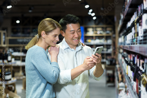 Young diverse couple man and woman in liquor department choosing wine  smiling and happy using smartphone to identify wine  using app to scan products