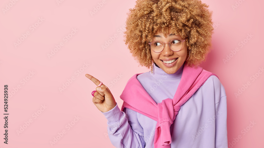 Horizontal shot of positive curly haired woman smiles happily points index finger away on blank space shows awesome advertisement wears transparent eyeglasses and casual jumper isolated on pink wall