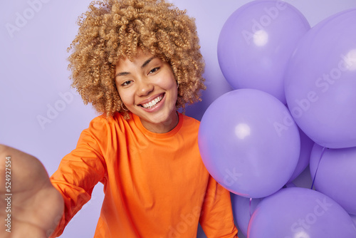 Happy curly haired woman stretches arm with unrecognizable device dressed in orange jumper makes selfie holds bunch of balloons celebrates special occasion smiles broadly isolated over purple wall