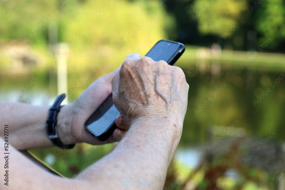 Elderly woman standing with smartphone in a park, mobile phone in wrinkled female hands closeup. Concept of online communication in retirement