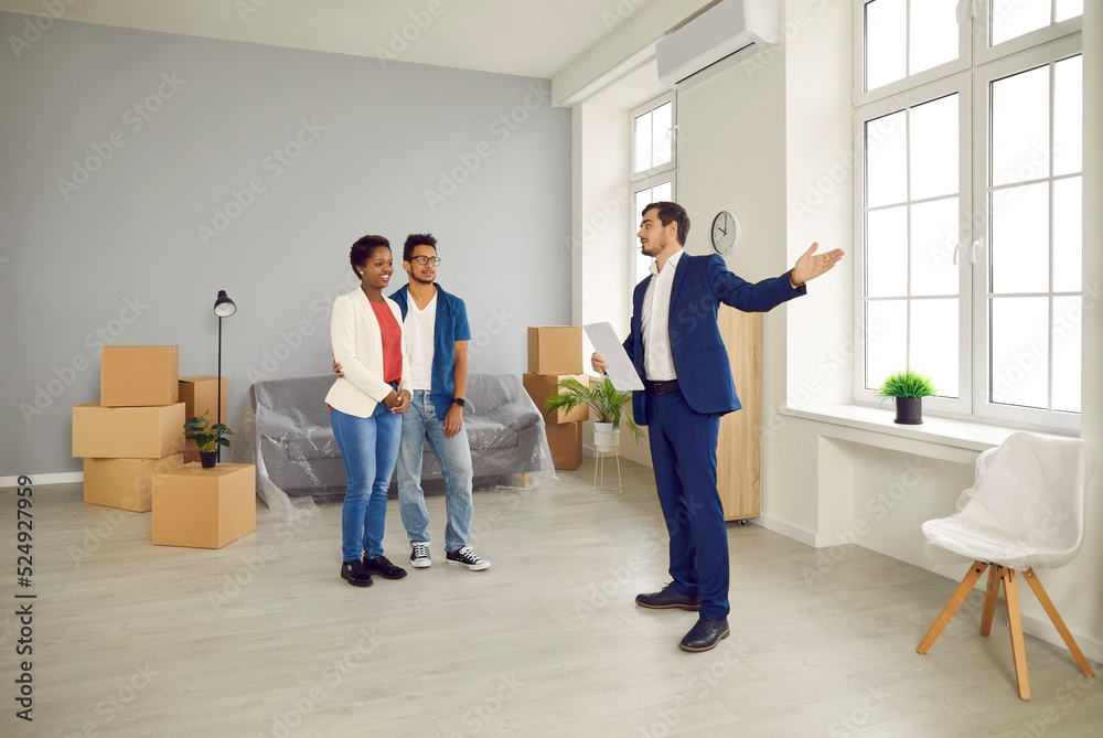 Fototapeta premium Real estate agent giving house tour to future tenants or potential buyers. Boyfriend and girlfriend or husband and wife planning to buy new home and meeting with realtor in modern, big, spacious flat