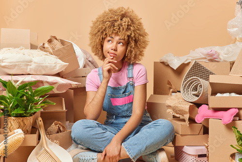Thoughtful curly haired young woman sits in lotus pose holds chin looks somewhere thinks about decor of her new apartment relocates to new place of living surrounded by stacks of cardboars boxes photo