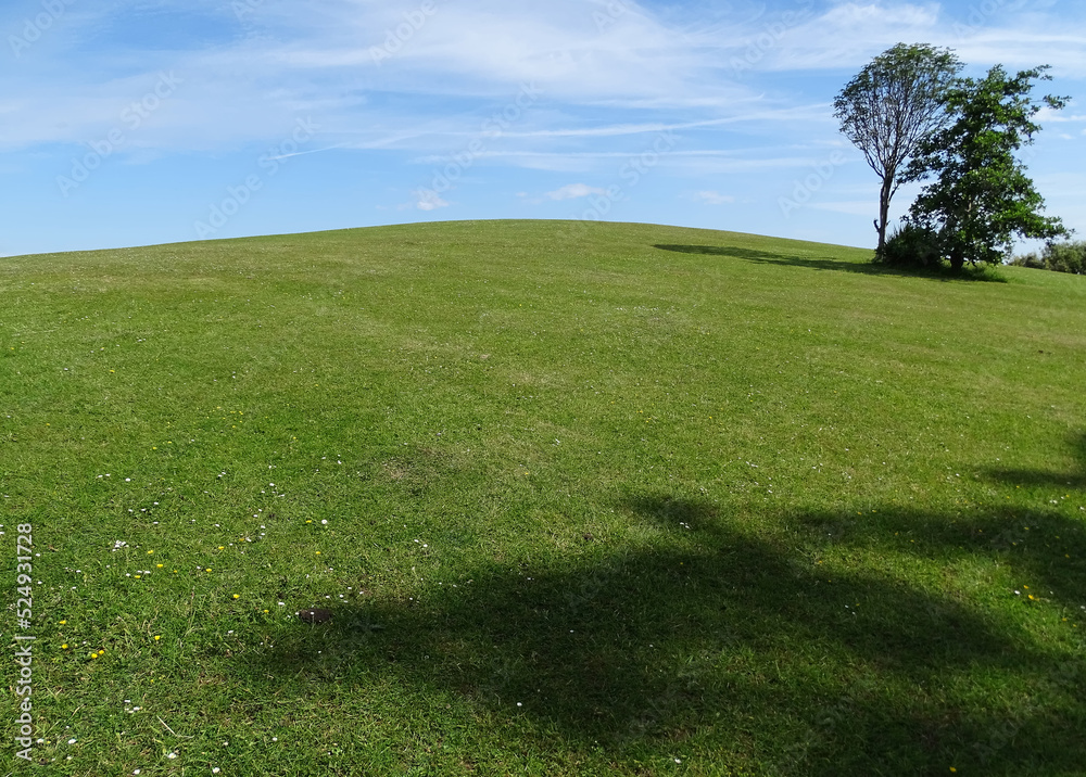 Green hill in foreground with sky in background. Light white clouds on a sunny day, vivid green grass with tree and shadows prominent taken in Colwyn Bay from Eirias Park Green hill in foreground with sky in background. Light white clouds on a sunny day, vivid green grass with tree and shadows prominent taken in Colwyn Bay from Eirias Park