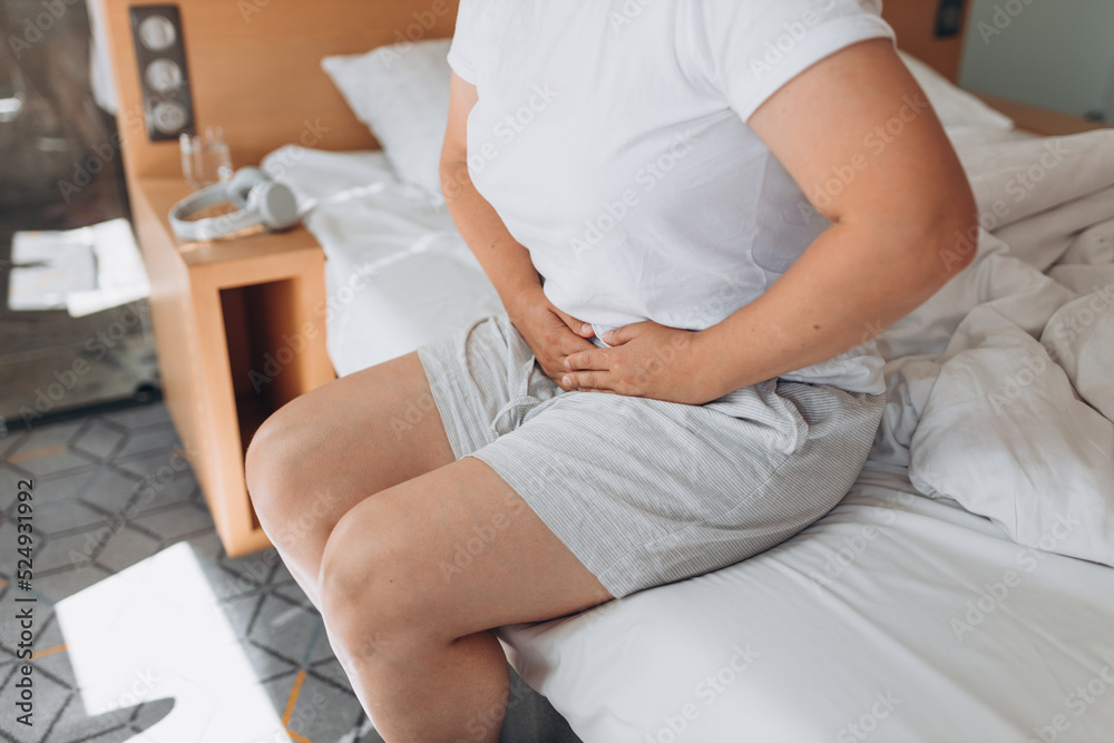 Young woman suffering from strong abdominal pain, casual style indoor shoot. Female sitting on bed in the morning and touching stomach. Sickness and Gynecology concept