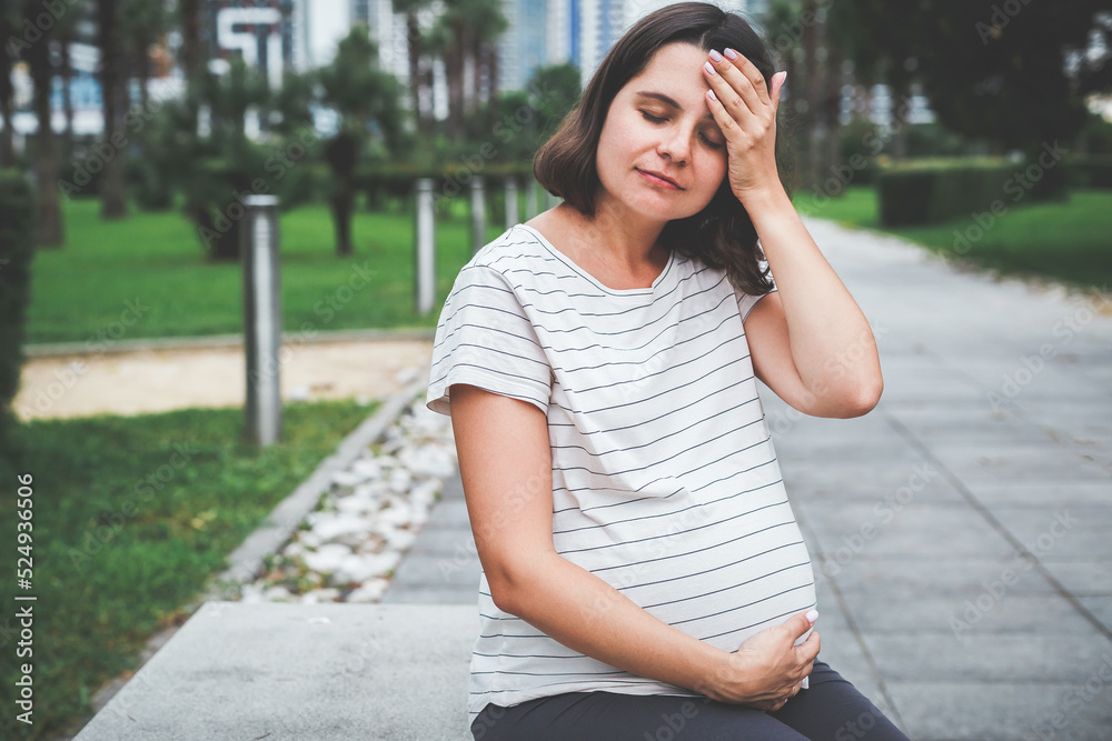 a sad pregnant woman sits alone on a bench in the park in summer holds on to her head and belly, discomfort and pain during pregnancy a sad pregnant woman sits alone on a bench in the park in summer holds on to her head and belly, discomfort and pain during pregnancy