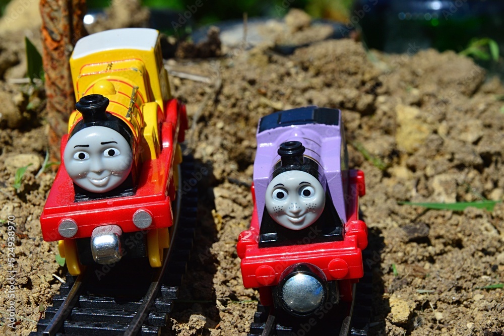 Toy cast iron train models of Molly (yellow) and Rosie (pink) from Thomas  The Tank Engine TV cartoon riding on HO model railway side-by-side on soil  surface in garden. Stock Photo