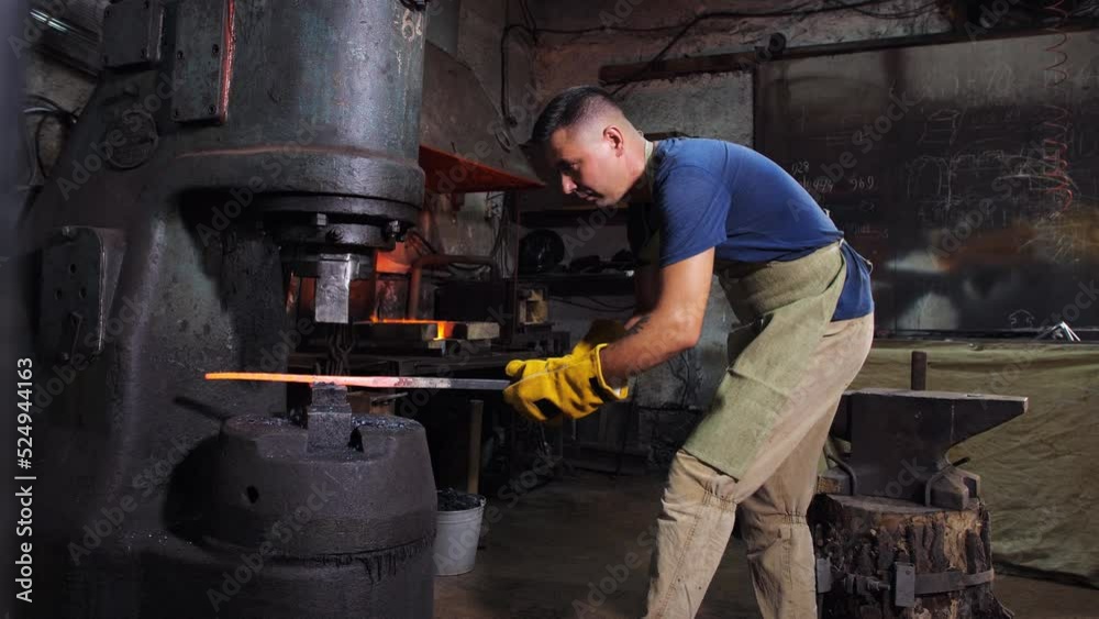 The process of working a mechanical anvil in a blacksmith shop. A blacksmith shapes a red-hot metal billet with a mechanical hammer. The process of working a mechanical anvil in a blacksmith shop. A blacksmith shapes a red-hot metal billet with a mechanical hammer.