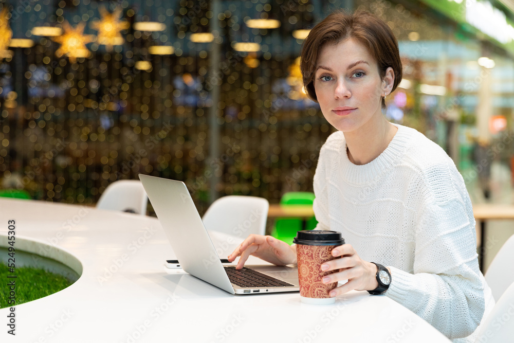 girl works on a laptop and drinks coffee with a smile looking at the camera