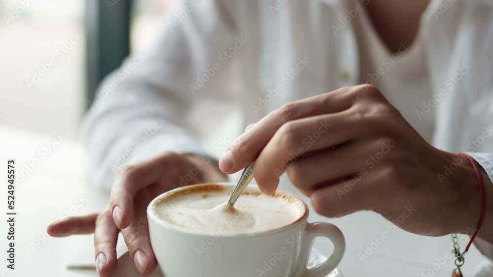 A hand stirs a cup of coffee with a spoon, cappuccino coffee in a white cup, stirring the coffee foam with a metal spoon. A girl drinks coffee sitting at a restaurant table Cappuccino with thick foam A hand stirs a cup of coffee with a spoon, cappuccino coffee in a white cup, stirring the coffee foam with a metal spoon. A girl drinks coffee sitting at a restaurant table Cappuccino with thick foam