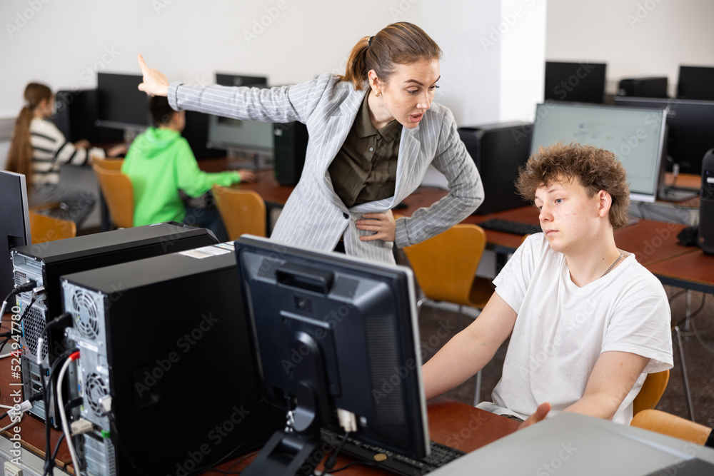 Strict young female teacher reprimanding negligent teenage student sitting at computer in classroom during lesson..