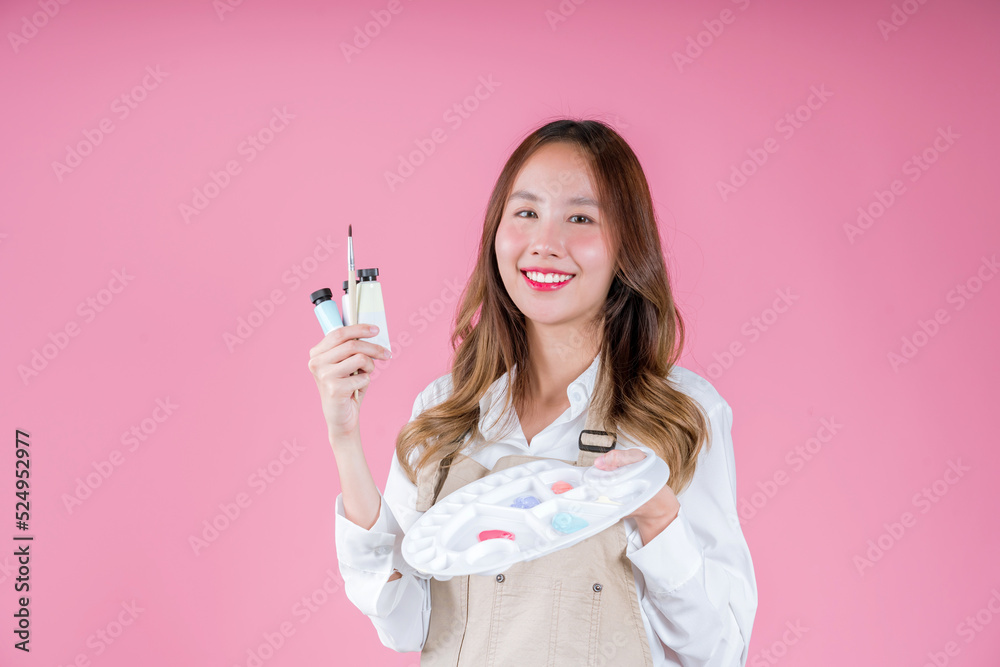 Beautiful Asian woman painter artist holding quill pen and color palette using paintbrush for desigh, drawing, paint workshop on pink background