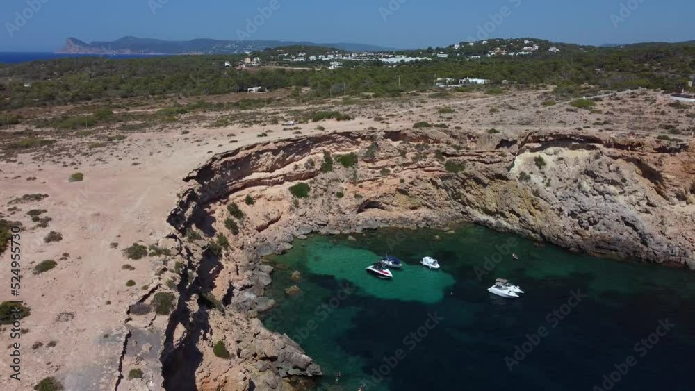 Emerald green bay rugged cliffs boats anchorDramatic aerial view flight pedestal down drone footageof cliff edge beach Sa Figuera borda Ibiza summer 2022