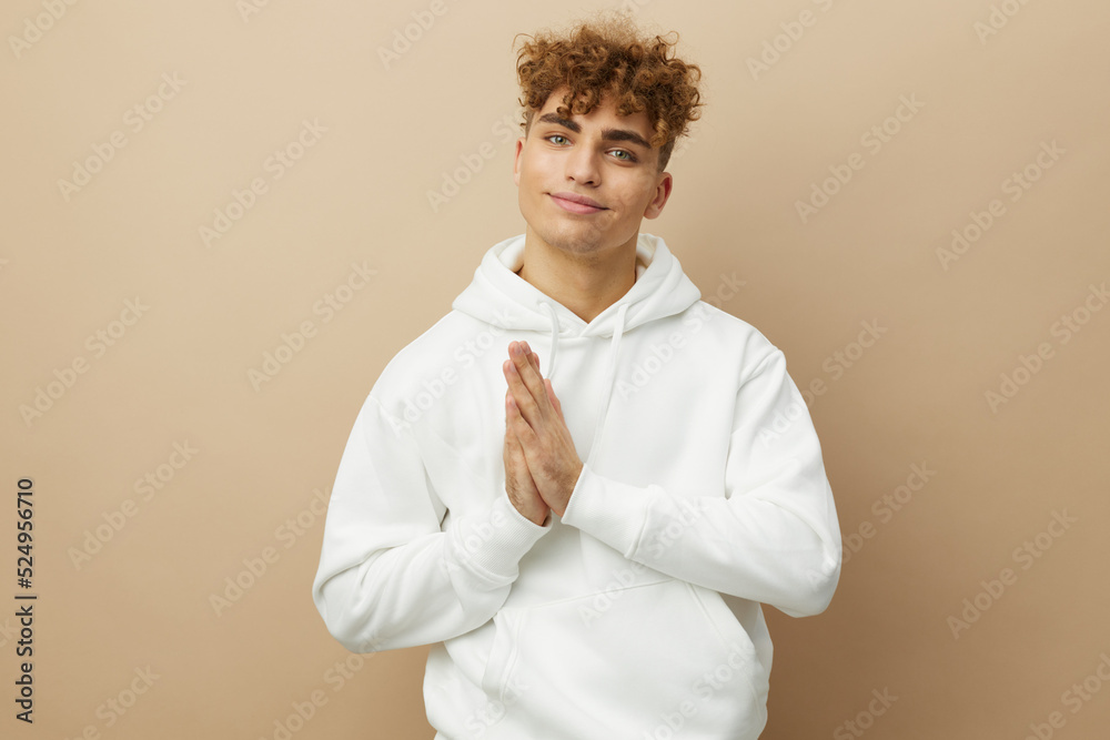 funny, happy man standing on a beige background in a white hoodie and smiling broadly holding his palms folded together funny, happy man standing on a beige background in a white hoodie and smiling broadly holding his palms folded together