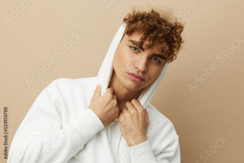 a stylish young man in a white hoodie, standing on a beige background and holding the hood on his head with his hands, smiling pleasantly at the camera
