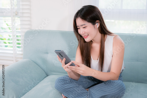 Asian woman use smartphone and wearing earbuds listening music entertainment her enjoy and fun sitting on sofa in living room at home