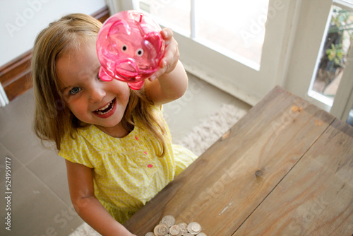 Smiling girl with blonde hair wearing yellow blouse raising a pink transparent piggy bank photo