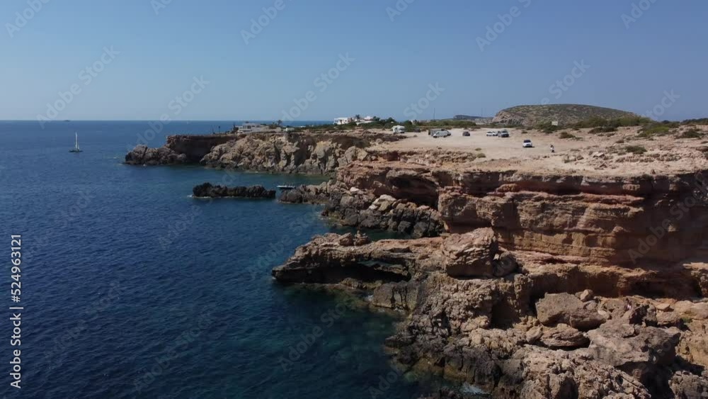 Sailboat in blue water, cars on rugged rock.Fantastic aerial view flight pedestal up drone footageof cliff edge beach Sa Figuera borda Ibiza summer 2022.