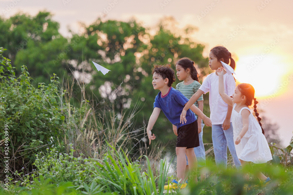 Fototapeta premium Cute asian little girl and her siblings playing with paper airplane toy together with fun. Group of asian kid throwing paper plane and playing together in the countryside at the sunset time.