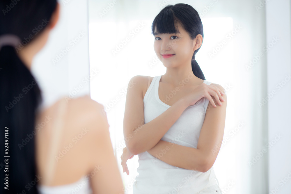 Beautiful young asian woman looking in mirror and apply moisturizer with cosmetic cream or lotion with shoulder in bathroom at home, skincare for hygiene, treatment and bodycare, skin care concepts.