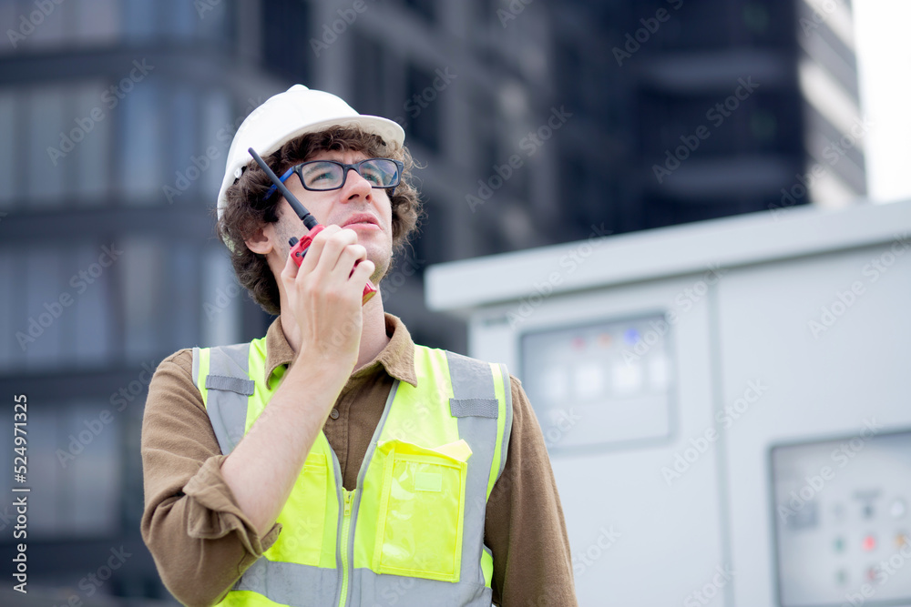 Fototapeta premium Young caucasian man using radio conversation and inspecting while building and tower all around, engineer or architect examining and maintenance for development, industrial and communication concept.