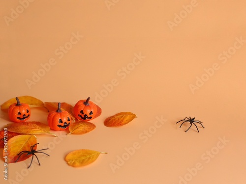 Three small Beautiful pumpkins, two black small spider and fallen yellow-red leaves on a orange background. Creative halloween concept backdrop. 