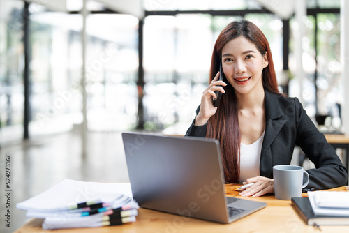 Happy businesswoman sitting at desk behind her laptop and talking with somebody on her mobile phone while working at office.