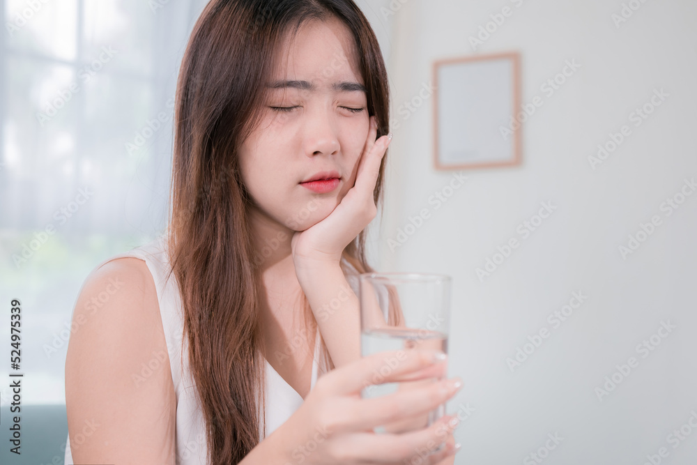 Asian woman has sensitive teeth after drinking cold water on sofa in living room at home. concept dental health Asian woman has sensitive teeth after drinking cold water on sofa in living room at home. concept dental health