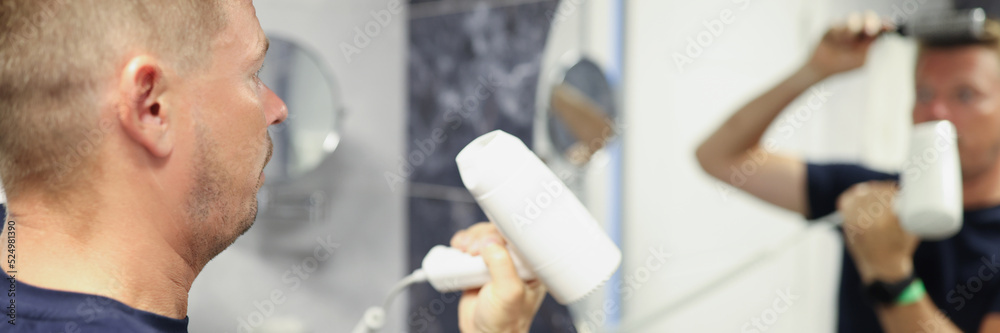 Man does hair styling with hairdryer and comb in front of mirror