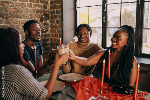 Best friends celebrating new year. Young people with candles, sitting at dining table. Diverse students during christmas party at home, smiling and laughing.