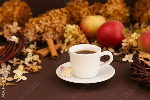 White coffee cup, autumn leaves, cinnamon, apples and dry hydrangea on brown background, front view. Selective focus