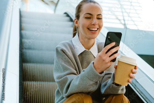 Young businesswoman holding coffee and cellphone sitting on escalator